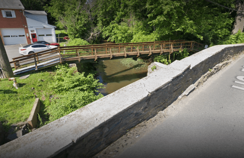 A wooden bridge over a small stream, surrounded by lush greenery, with a road and a car visible nearby.