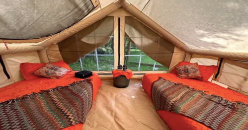 Cozy glamping tent interior with two orange beds, decorative pillows, and a view of greenery outside.