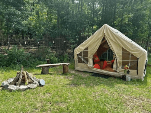 A cozy tent in a forest clearing, with a campfire setup and a wooden bench nearby, surrounded by greenery.