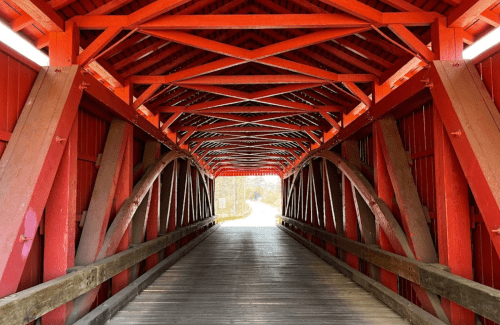 A view inside a red covered bridge, showcasing wooden beams and a pathway leading to the outside.