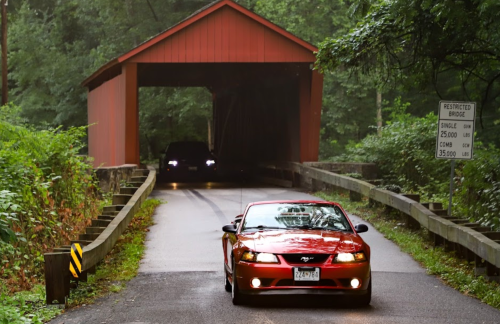 A red car drives through a covered bridge surrounded by lush greenery. Another vehicle approaches from the opposite direction.