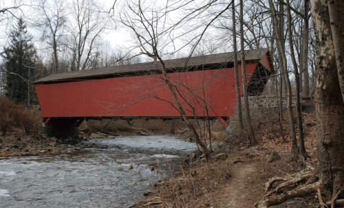 A red covered bridge spans a stream, surrounded by bare trees and a rocky bank in a tranquil, wooded setting.