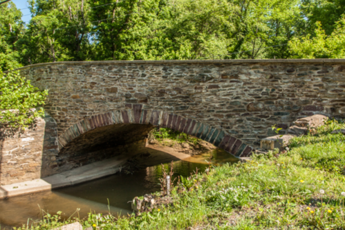 A stone arch bridge over a small stream, surrounded by lush greenery and trees.