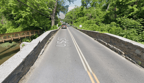 A road bridge over a river, surrounded by lush green trees, with cars driving on the asphalt.