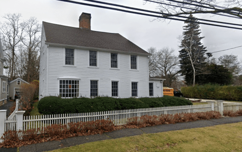 A white two-story house with a front yard, surrounded by a white picket fence, on a cloudy day.