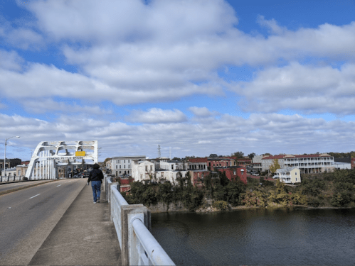 A person walks along a bridge with a view of a river and colorful buildings under a partly cloudy sky.
