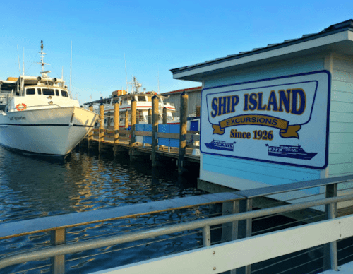 A dock with boats and a sign for Ship Island Excursions, established in 1926, by the water.