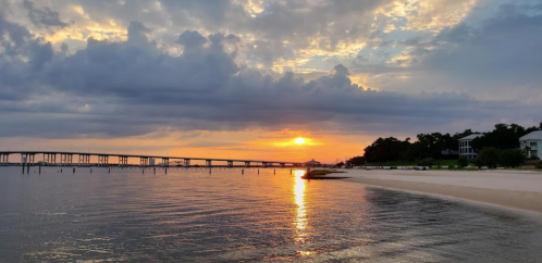 Sunset over calm waters with a bridge in the distance and a sandy beach lined with trees and houses.