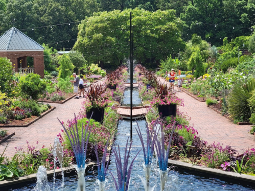 A vibrant garden with a central water feature, surrounded by colorful flowers and visitors enjoying the scenery.