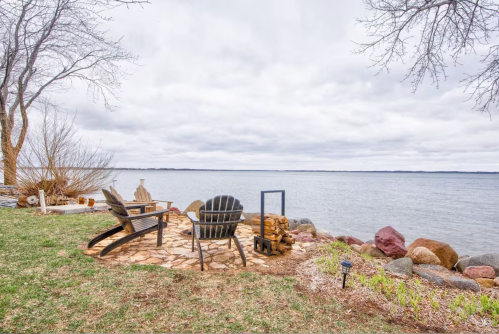 A serene lakeside scene with two chairs on a stone patio, surrounded by grass and rocks under a cloudy sky.