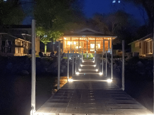 A lit wooden dock leads to a cozy house at night, surrounded by trees and water.