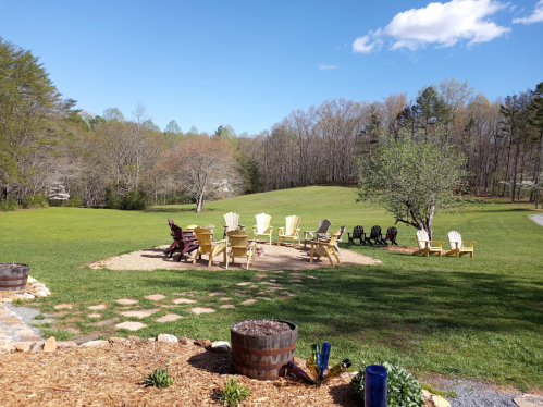 A scenic outdoor area with wooden chairs arranged in a circle on a grassy field, surrounded by trees and blue sky.