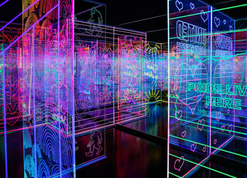 Colorful neon light installations with vibrant patterns and messages celebrating pride in a dark space.
