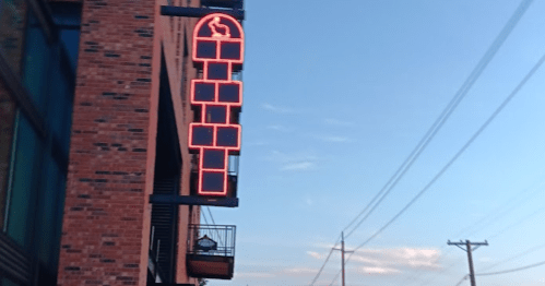 A brick building with a large neon sign, street view with parked cars and trees under a clear sky.