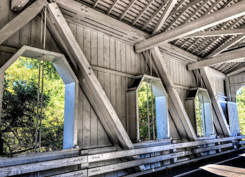 Interior view of a wooden covered bridge, featuring exposed beams and large windows with greenery outside.