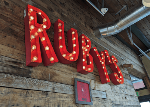 Bright red "RUBY'S" sign with light bulbs, mounted on a rustic wooden wall.