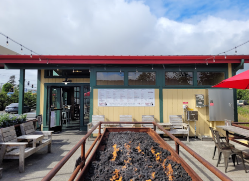 A restaurant exterior with a red roof, outdoor seating, and a fire pit in front, featuring a menu on the wall.
