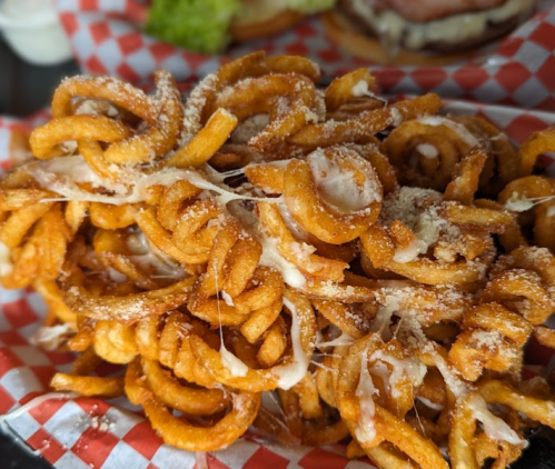 A close-up of crispy, golden curly fries topped with melted cheese, served in a red and white checkered basket.