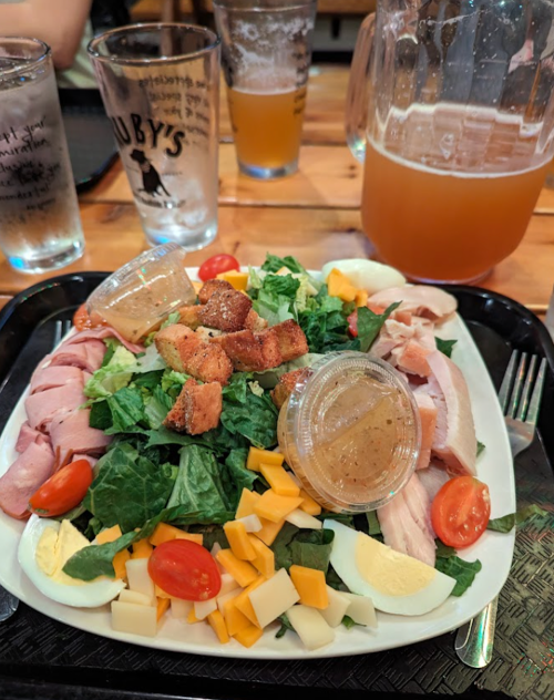 A colorful salad with greens, tomatoes, cheese, turkey, ham, croutons, and dressing, alongside drinks in glasses.