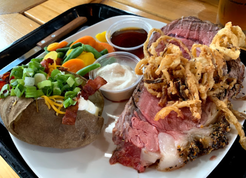A plate featuring prime rib, crispy onions, a loaded baked potato, and mixed vegetables with dipping sauces.