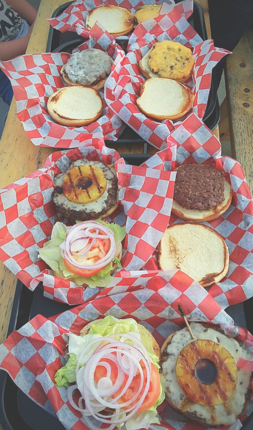 A variety of burgers on trays with red and white checkered paper, some with toppings and others without buns.