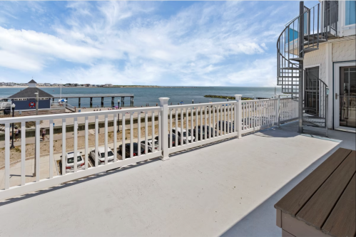 View from a balcony overlooking a beach, pier, and water, with a spiral staircase and parked cars below.