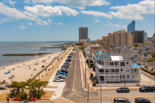 A coastal view featuring a beach, boardwalk, and city skyline under a clear blue sky with scattered clouds.