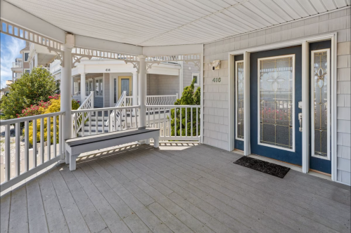 A covered porch with a bench and double doors, featuring a light-colored wooden floor and decorative railings.