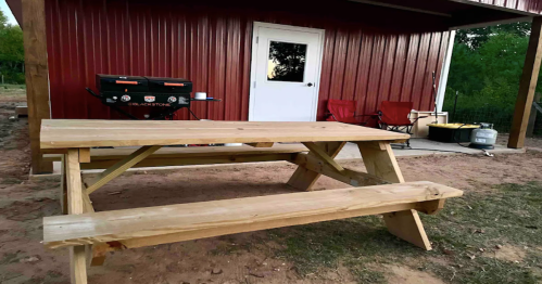 A wooden picnic table in front of a red shed, with a grill and two chairs nearby.