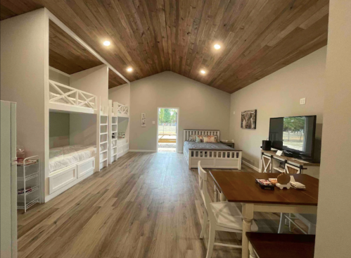 Bright, spacious room with wooden ceiling, bunk beds, a bed, TV, and dining table. Natural light from the door.