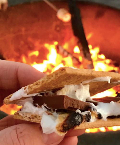 A hand holding a s'more with melted marshmallow and chocolate, with a campfire glowing in the background.
