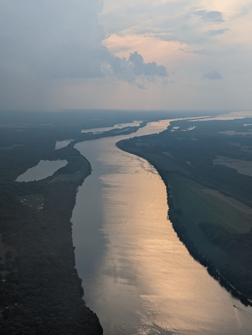 Aerial view of a winding river reflecting sunlight, surrounded by lush greenery and fields under a cloudy sky.