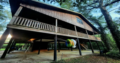 A wooden house on stilts surrounded by trees, featuring a spacious porch and colorful outdoor equipment below.