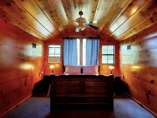 Cozy bedroom with wooden walls, a ceiling fan, and a bed flanked by lamps, featuring large windows with curtains.