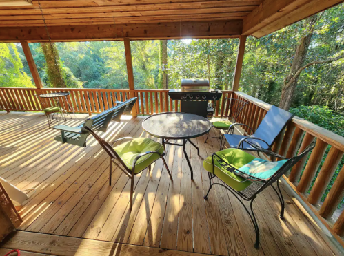 A cozy porch with wooden flooring, a grill, and seating surrounded by trees and sunlight.