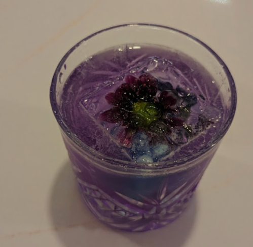 A purple drink with a floral ice cube, served in a textured glass, resting on a light surface.