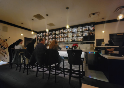 A cozy bar scene with patrons seated at the counter, illuminated by warm lights and a wall of bottles behind them.