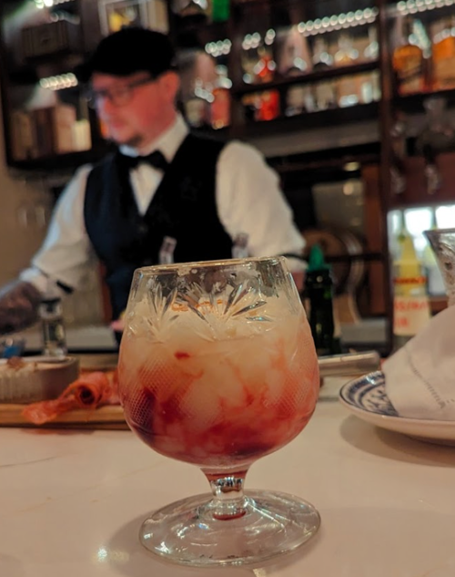 A close-up of a cocktail in a glass, with a blurred bartender in the background.
