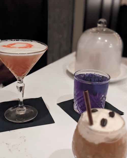 Three colorful cocktails on a table: a pink martini, a blue drink, and a creamy brown beverage with coffee beans.