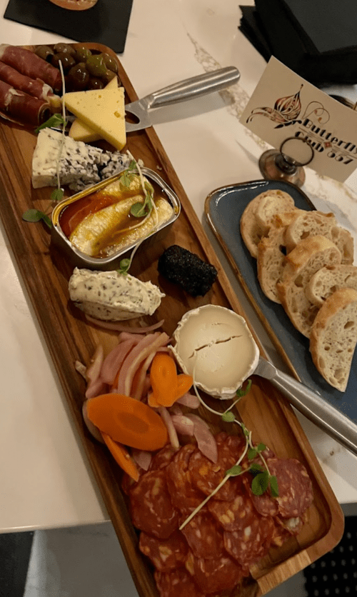A wooden platter featuring assorted cheeses, cured meats, olives, pickled vegetables, and slices of bread.