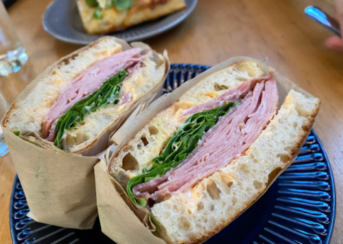 A close-up of a sandwich with layers of ham and greens, served on a blue plate with a side dish in the background.