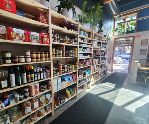 A well-stocked grocery store aisle with shelves of jars, bottles, and packaged goods, illuminated by natural light.