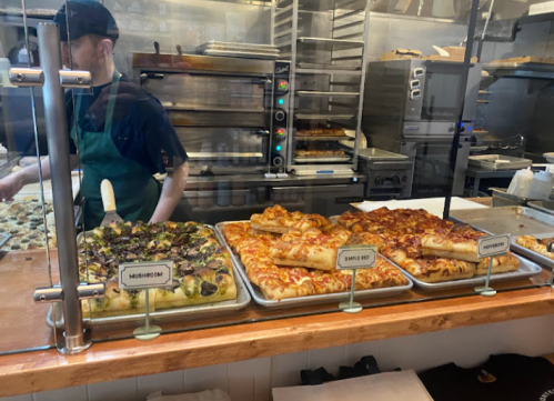 A display of various pizzas, including mushroom and simple flat, in a restaurant kitchen with a staff member in the background.