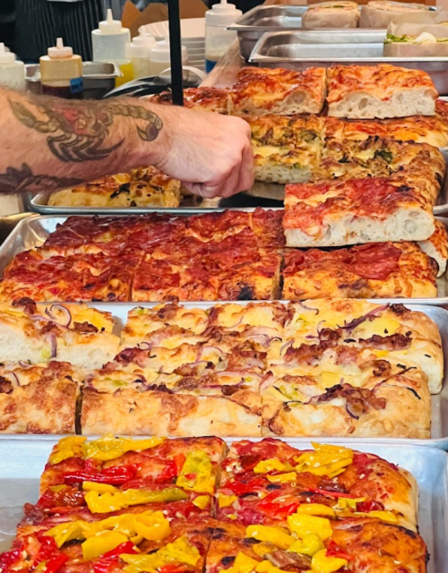 A hand reaches for a slice of colorful, freshly baked pizza on a tray, surrounded by various pizza styles.