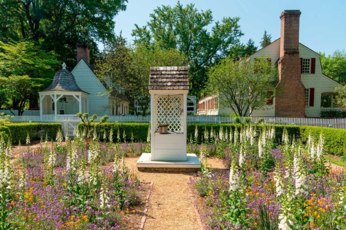 A charming garden with colorful flowers, a white gazebo, and a small well, surrounded by greenery and historic buildings.