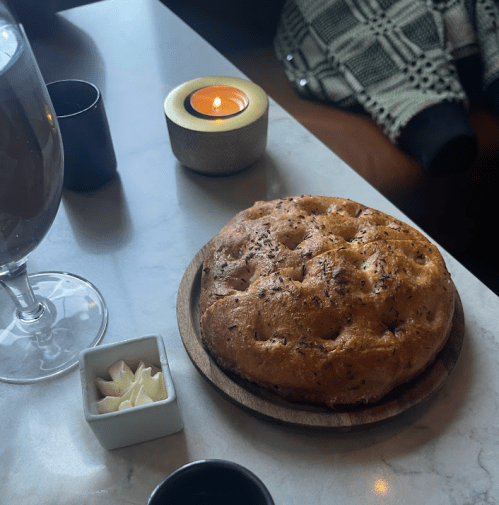 A wooden plate with a round, crusty bread, a small dish of butter, a glass of drink, and a lit candle on a table.