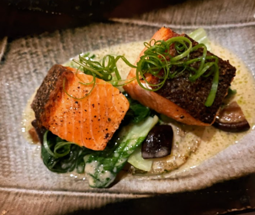Two pieces of seared salmon on a bed of greens, drizzled with sauce and garnished with green onions.