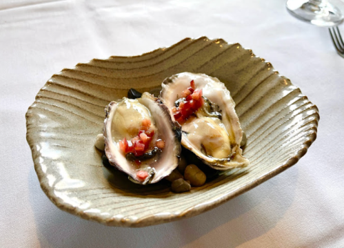 Two fresh oysters on a decorative plate, topped with diced tomatoes and served on a textured surface.
