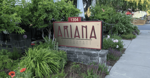 Sign for Ariana Restaurant surrounded by lush greenery and colorful flowers, located at 1304.