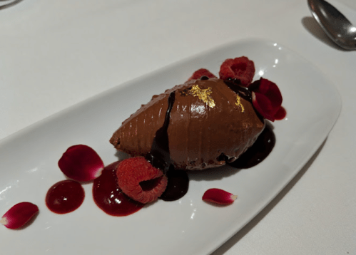 A chocolate mousse topped with gold leaf, surrounded by raspberry sauce and rose petals on a white plate.